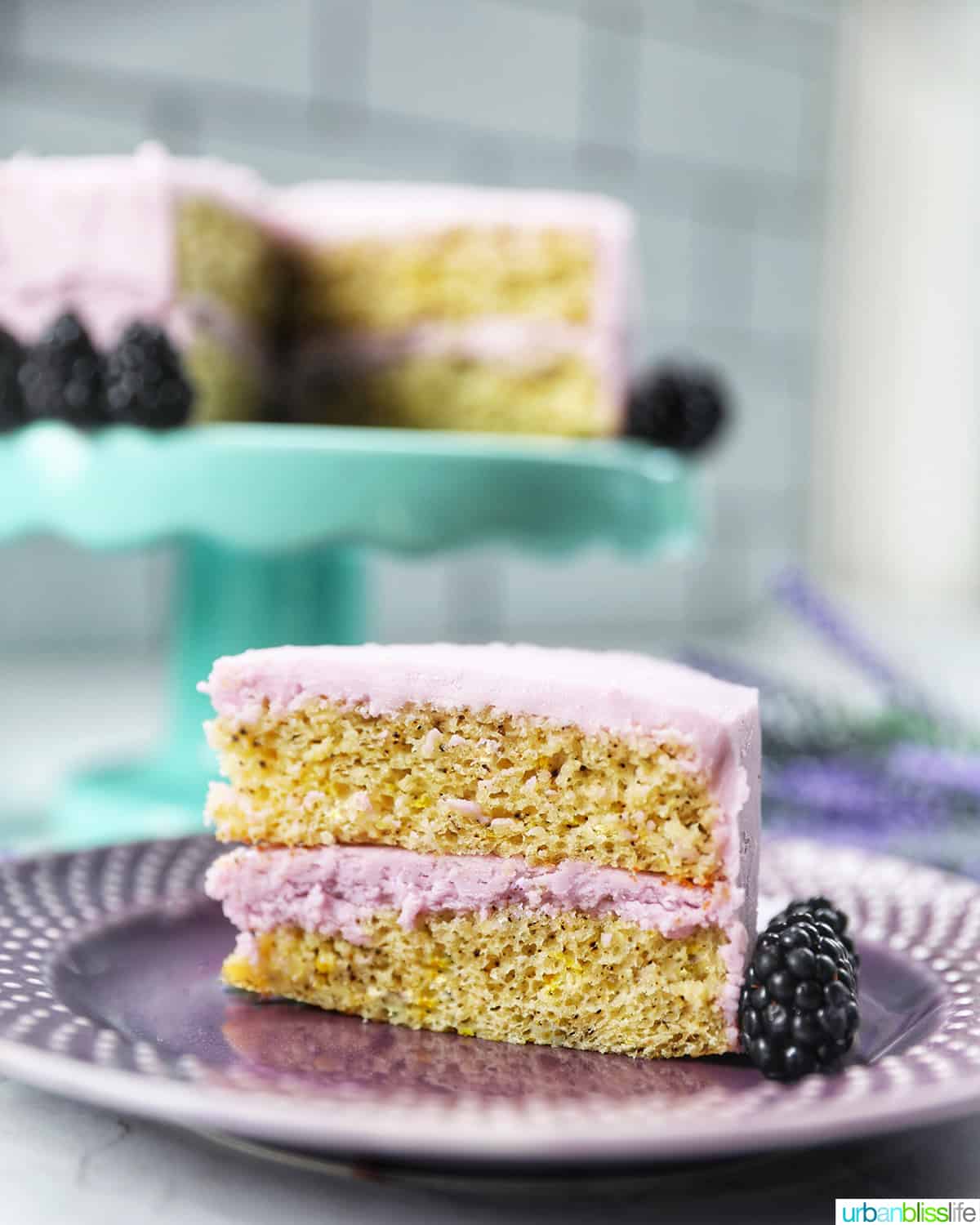 slice of earl grey lavender cake on a purple plate with blackberries with cake on an aqua cake pedestal in the background.