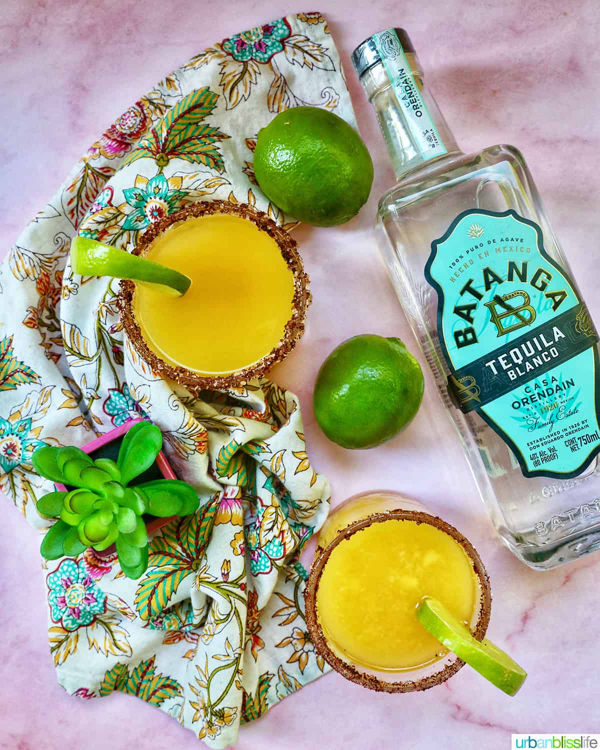 two glasses of spicy mango margarita with lime slice garnish and chili powder salt rim and bottle of tequila blanco.