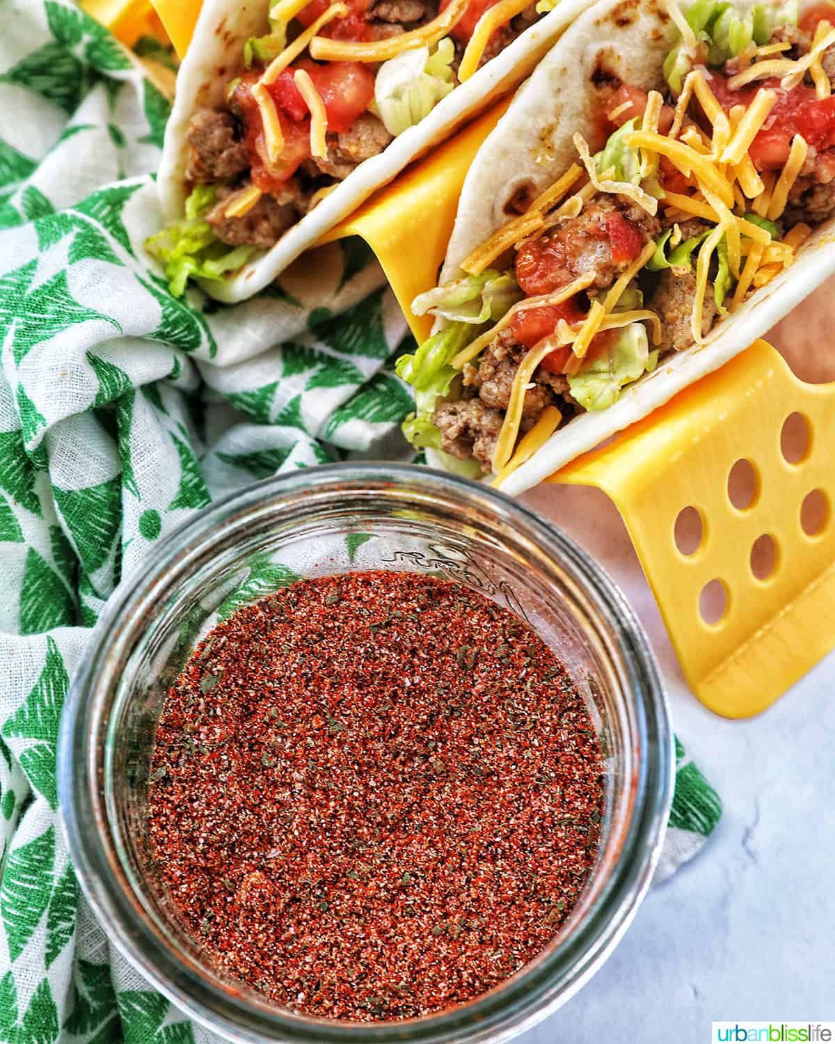 two tacos in a taco stand next to a jar of homemade mild taco seasoning with green and white napkin.
