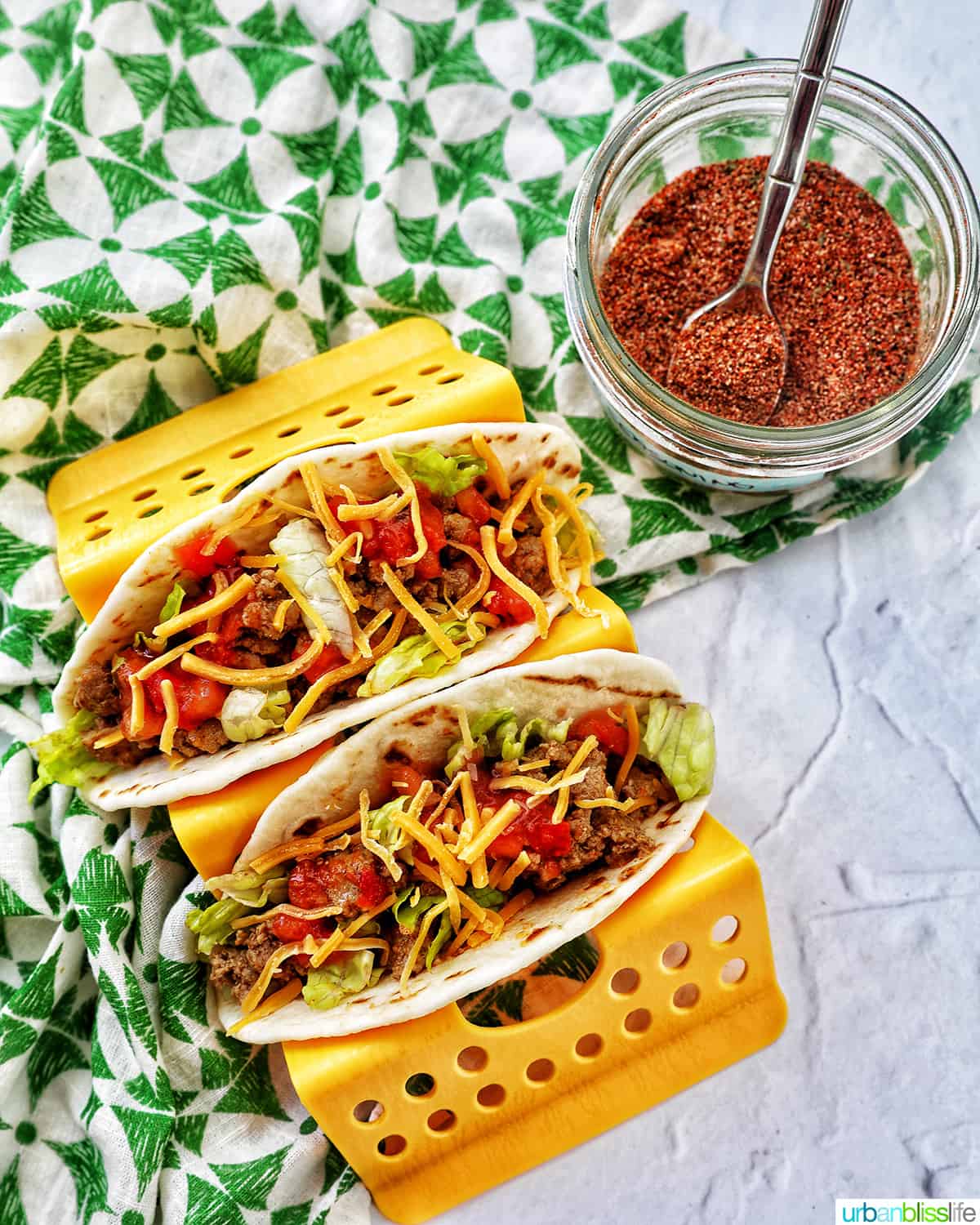 two tacos in a taco stand next to a jar of homemade mild taco seasoning with green and white napkin.