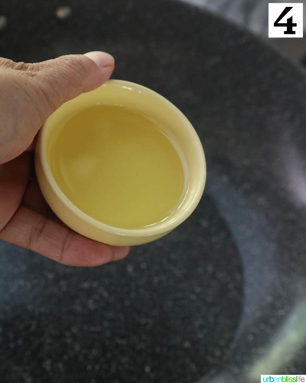 hand holding a bowl of olive oil over a wok.