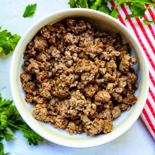 cooked ground beef in a white bowl with parsley sprigs around it and a red and white striped towel.