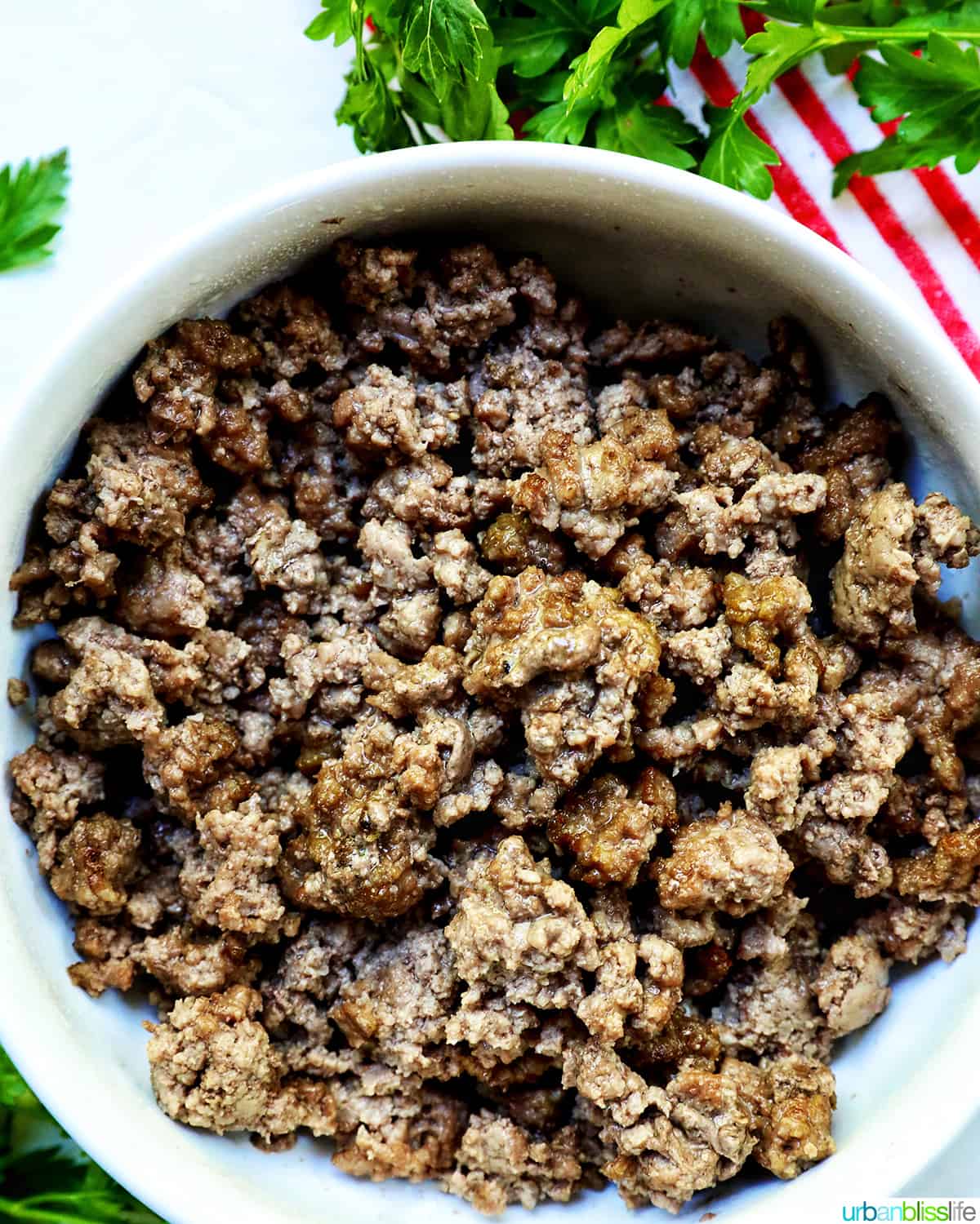 cooked ground beef in a white bowl with parsley sprigs around it and a red and white striped towel.