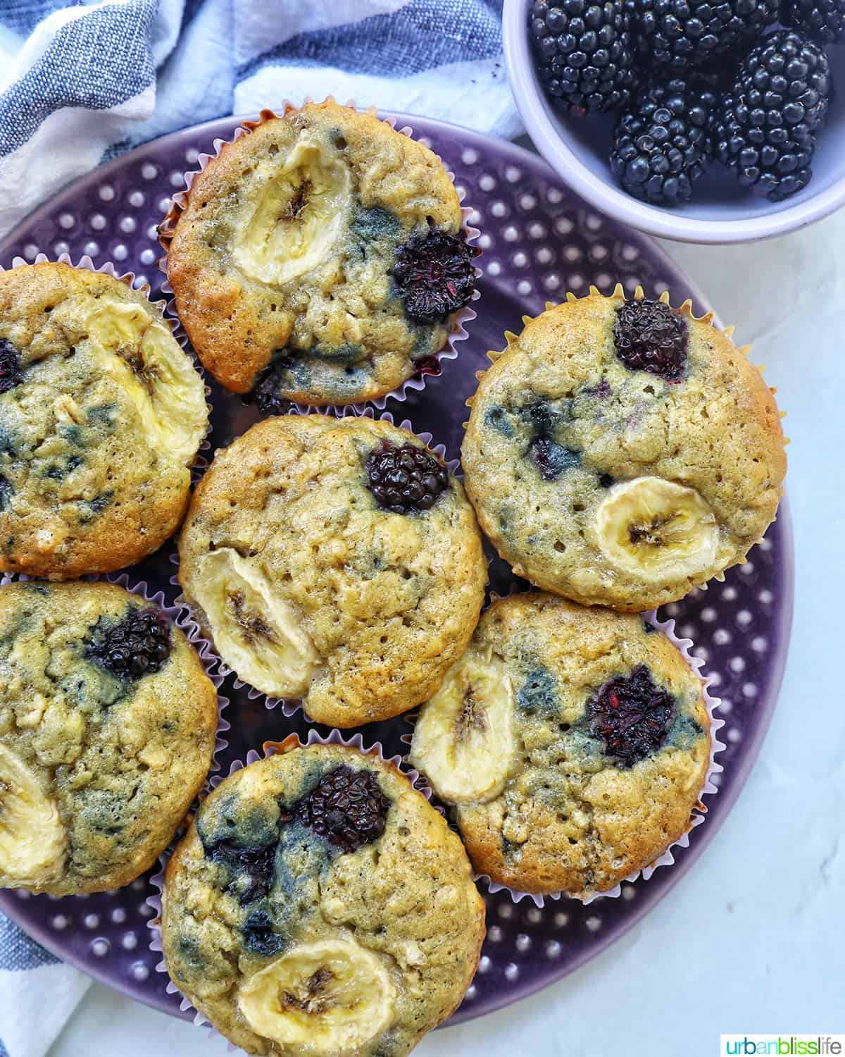 purple plate with several banana blackberry oatmeal muffins on top next to a side of blackberries.