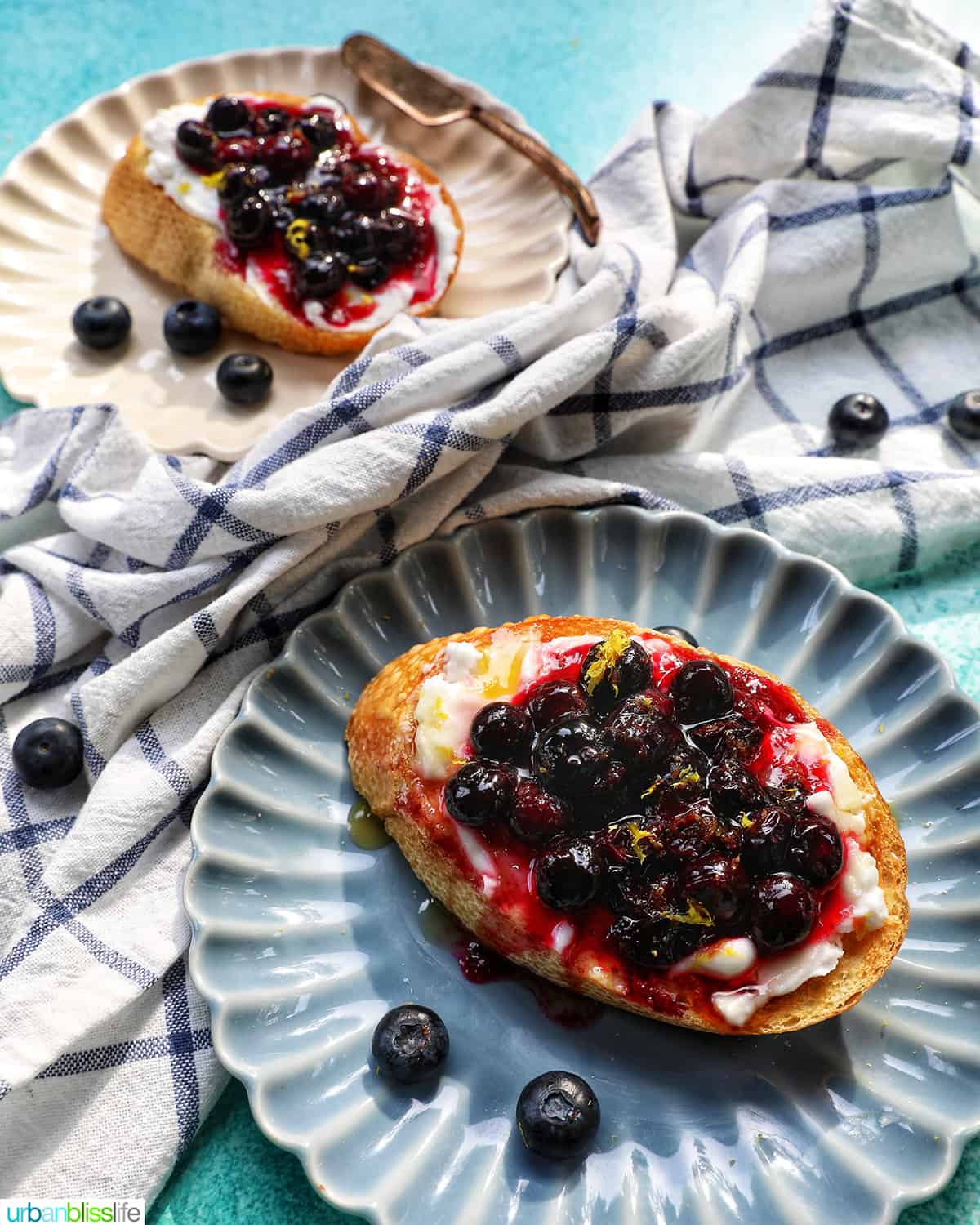 blueberries and cream cheese on toast on a blue scalloped plate, with a white plate of another blueberry toast in the background.