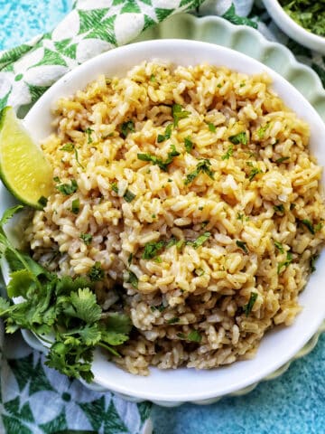 white bowl with Instant Pot Cilantro Lime Brown Rice on a green and white napkin with sides of cilantro leaves and lime wedges.