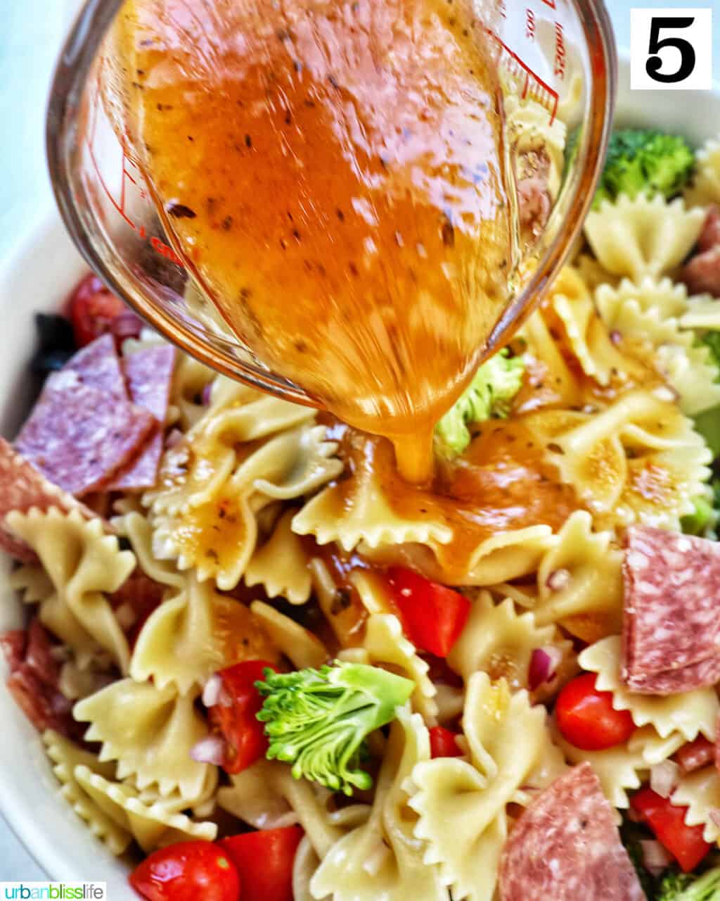 Italian dressing being poured over a bowl of Italian farfalle pasta salad.