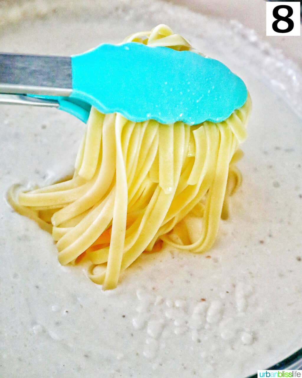 blue tongs holding pasta over cream sauce in a large skillet.
