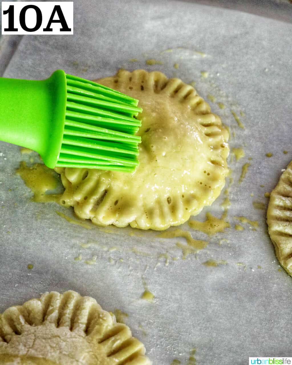 green pastry brush brushing egg wash onto a mini peach mango hand pie.