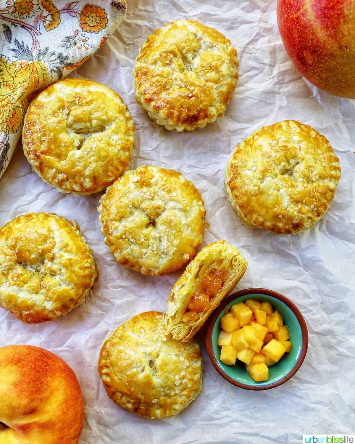 several mini peach mango hand pies on crinkled parchment paper with a bowl of diced peaches and mangoes.