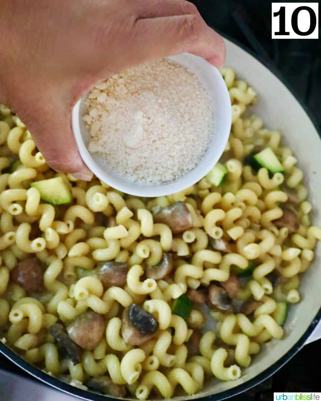 hand holding a bowl of grated parmesan cheese over mushroom zucchini pasta.