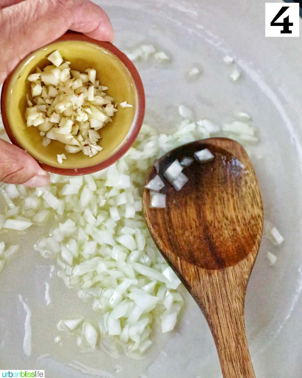 hand holding a bowl of minced garlic over a pan with onions, olive oil, and a wooden spoon.