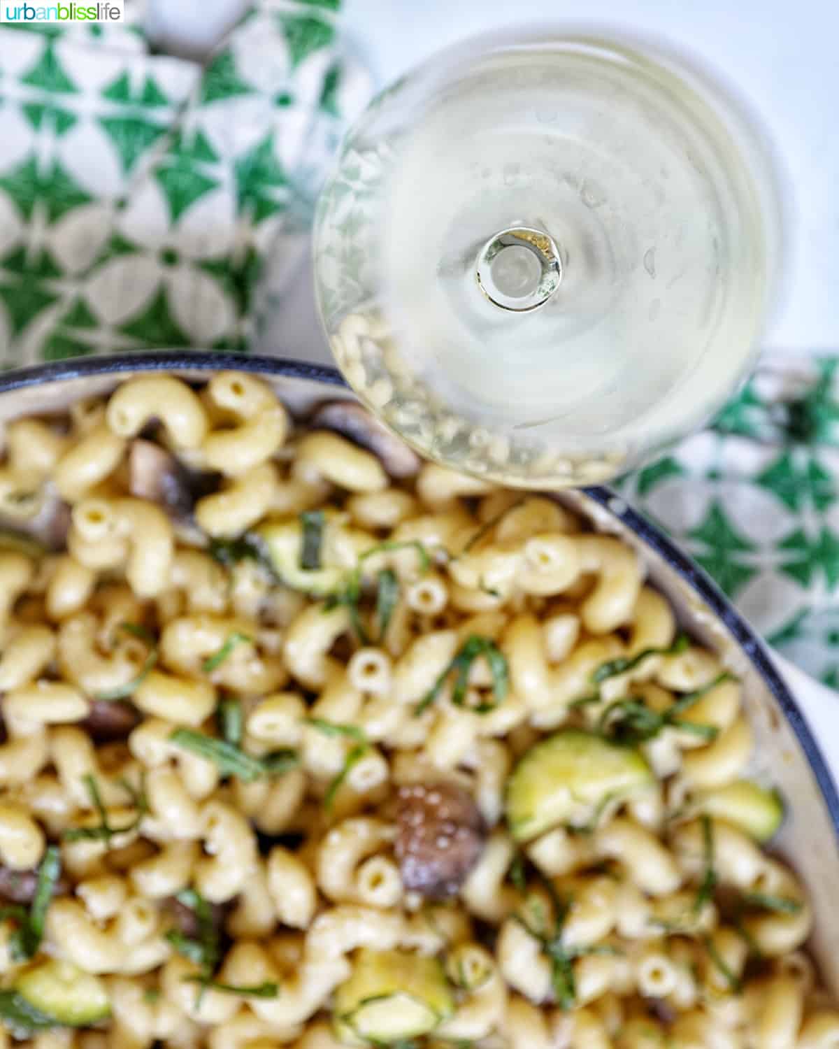 overhead photo of a glass of white wine next to pan of pasta with a green and white napkin.