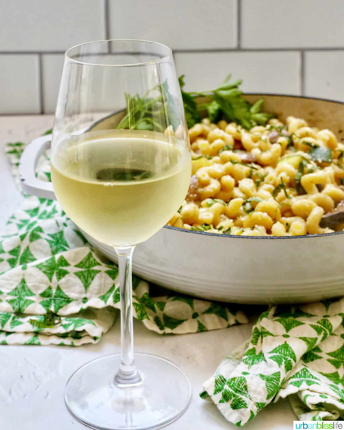 glass of white wine in front of a pan of pasta with a green and white napkin.