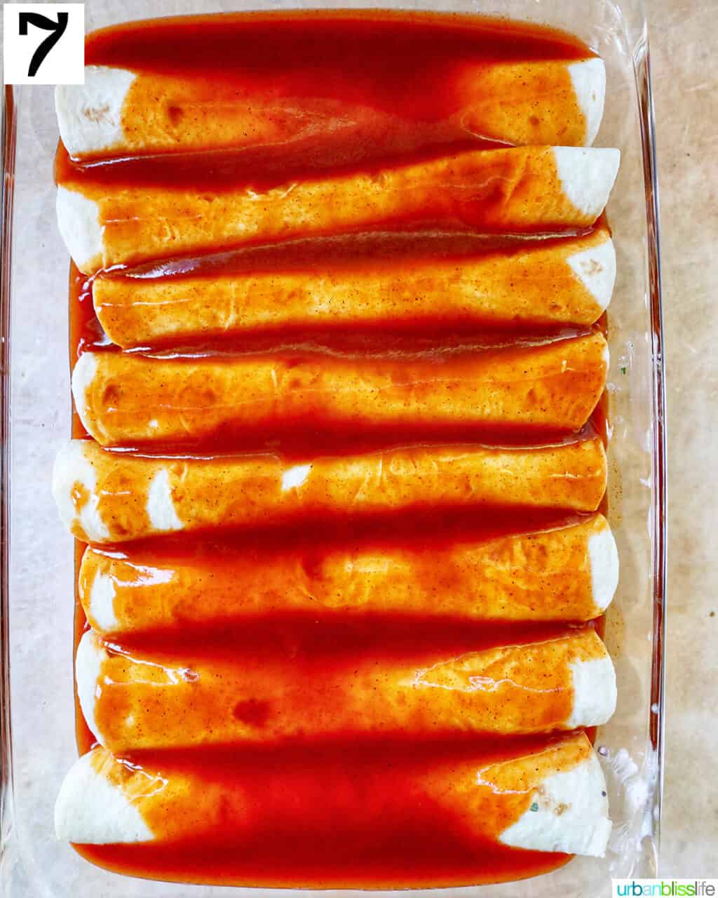 enchilada sauce over rolled flour tortilla enchiladas in a glass baking dish.