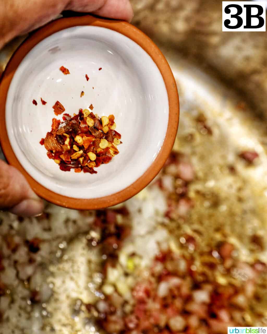 red pepper flakes in a small bowl above a saucepan with garlic, onions, and bacon.