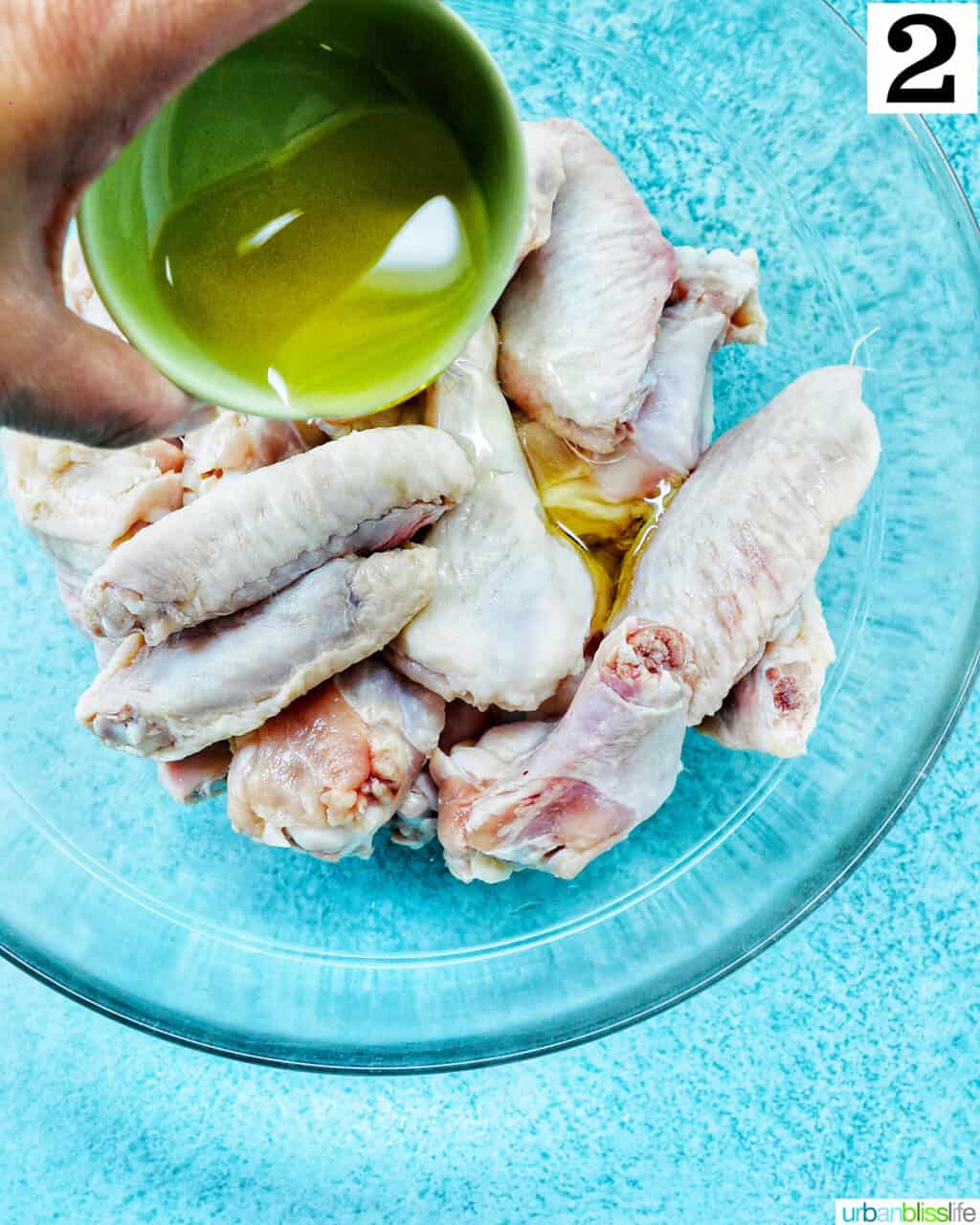 hand pouring olive oil over raw chicken wings in a large glass bowl.