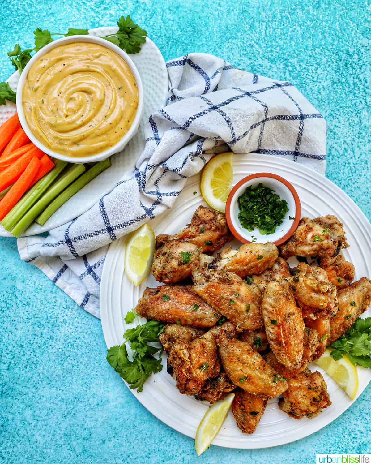 white plate full of salt and pepper wings with side of chopped fresh parsley and lemon wedges, and bowl of buffalo ranch sauce with carrots and celery.