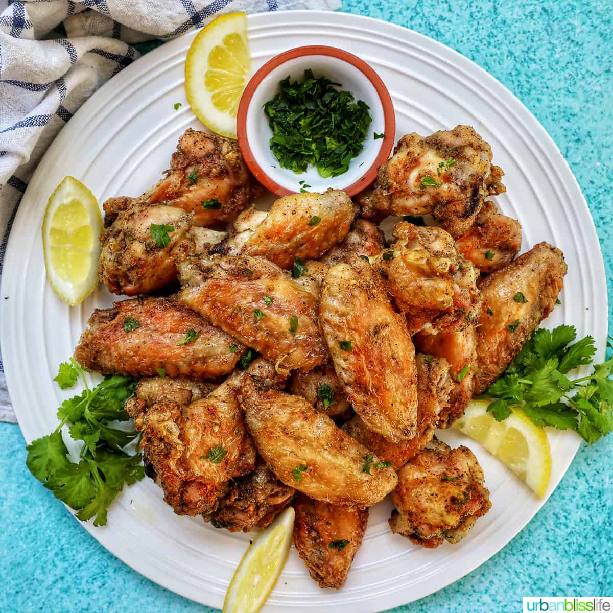 white plate full of salt and pepper wings with side of chopped fresh parsley and lemon wedges.