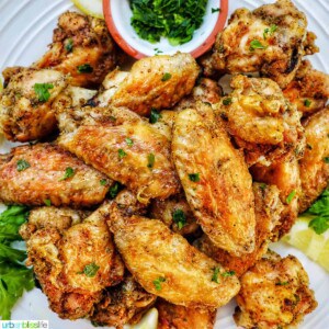 white plate full of salt and pepper wings with side of chopped fresh parsley and lemon wedges.