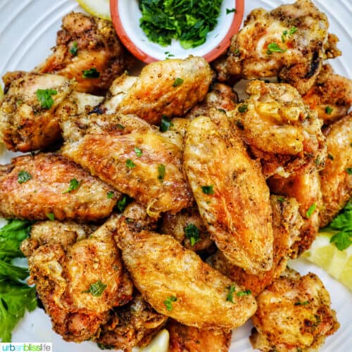 white plate full of salt and pepper wings with side of chopped fresh parsley and lemon wedges.