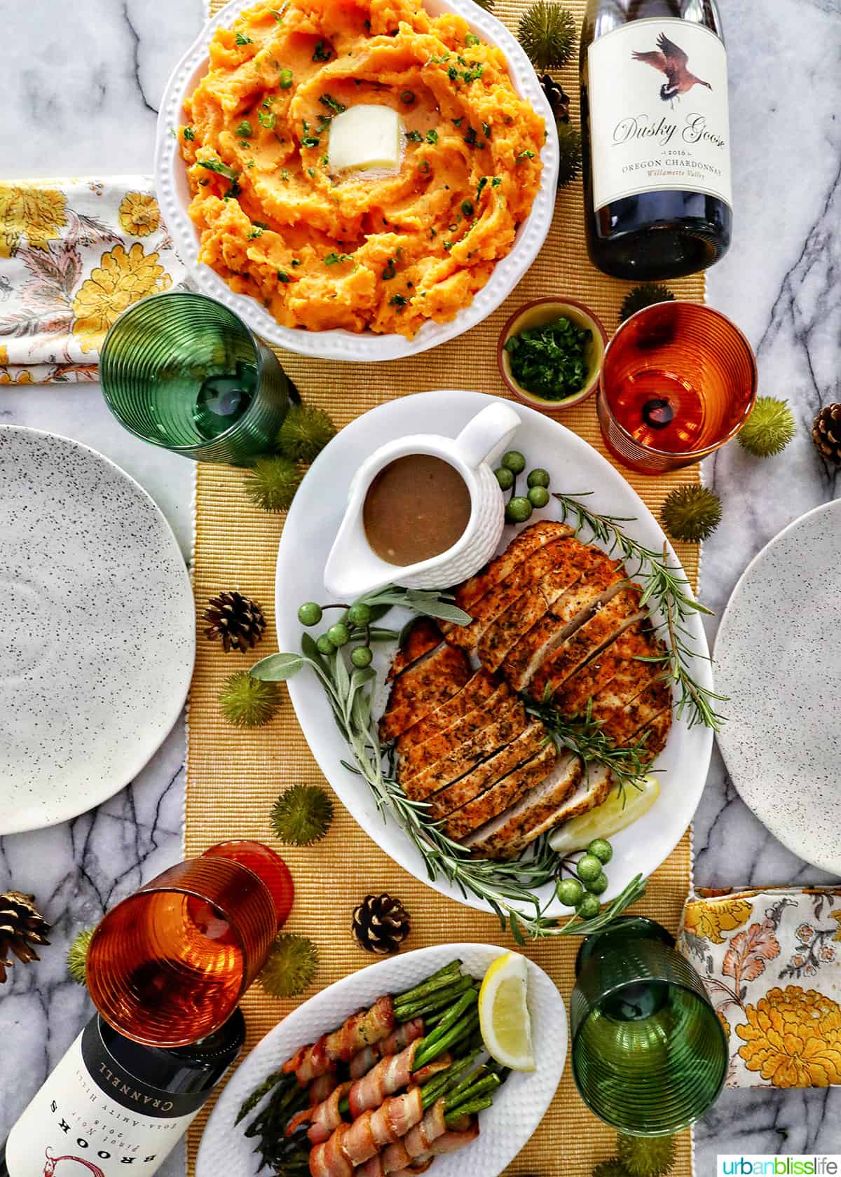 Thanksgiving dinner table with sliced turkey tenderloin, whipped sweet potatoes, bacon wrapped asparagus, bottles of wine, and servingware.