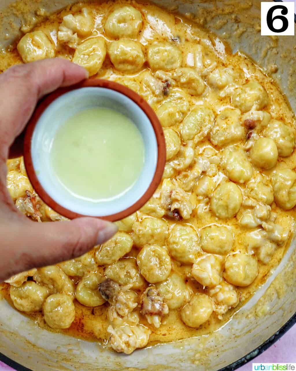 hand holding a bowl of lemon juice over gnocchi in a skillet.