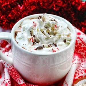 white mug with peppermint hot chocolate, whipped cream, surrounded by red and white Christmas decorations.