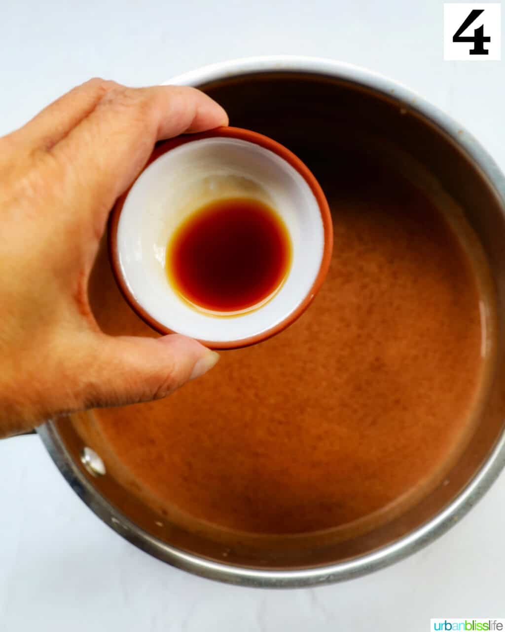 hand holding a bowl of vanilla extract over hot chocolate in a saucepan.