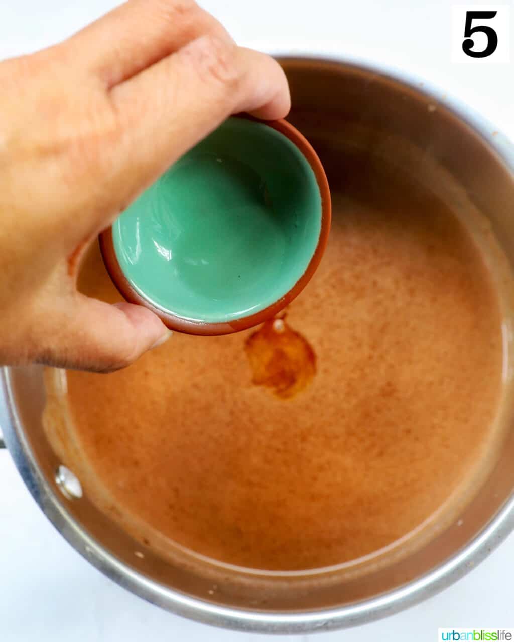hand holding a bowl of peppermint extract over hot chocolate in a saucepan.