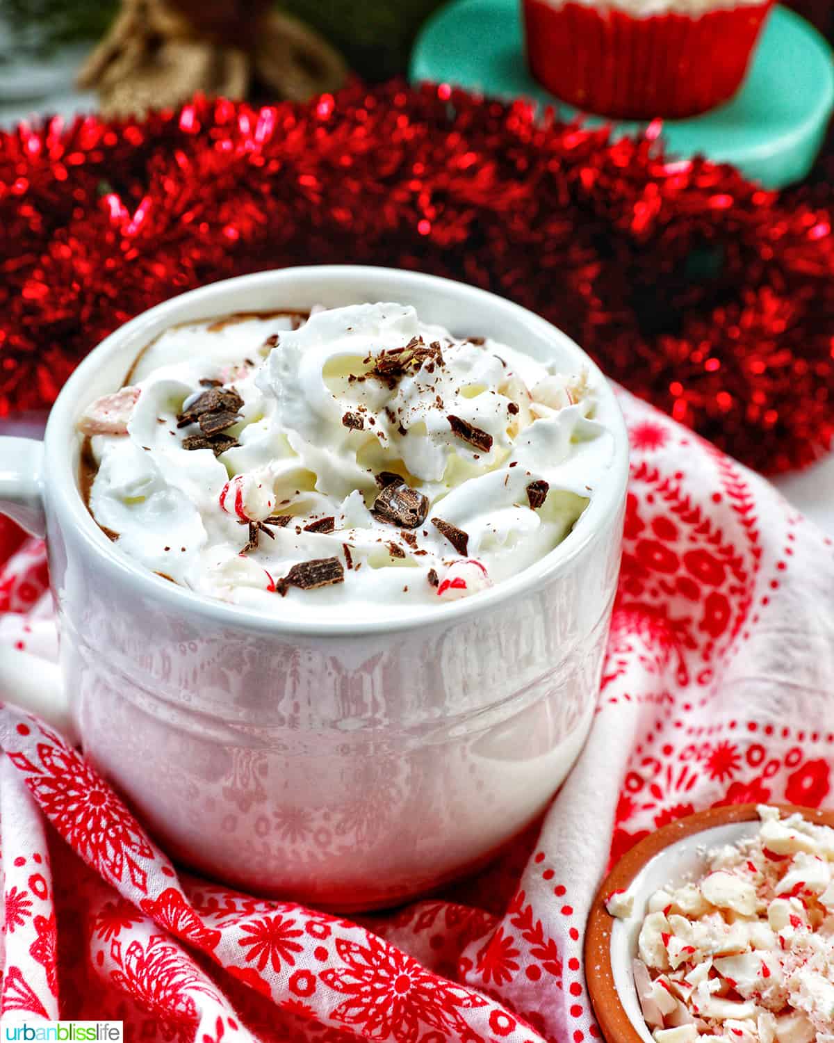 white mug with peppermint hot chocolate, whipped cream, surrounded by red and white Christmas decorations.
