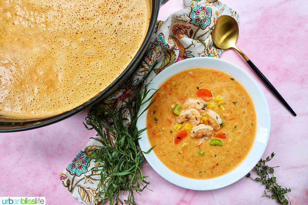 dutch oven full of soup next to a bowl of shrimp and corn bisque with fresh herbs and a gold spoon on pink table.