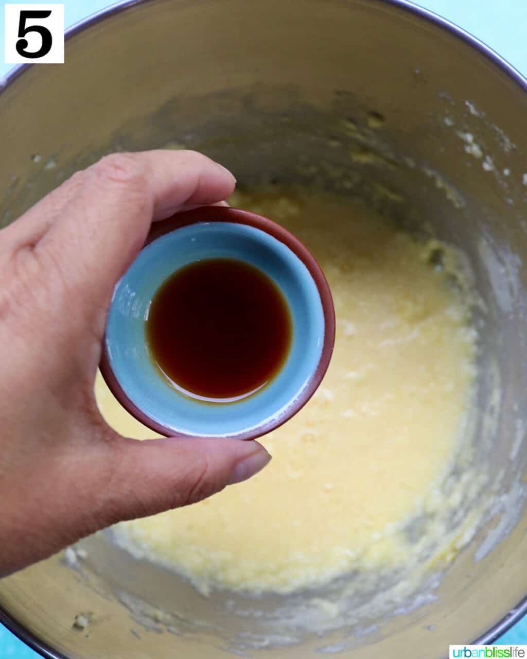 hand holding vanilla extract over cupcake batter in stainless steel bowl.