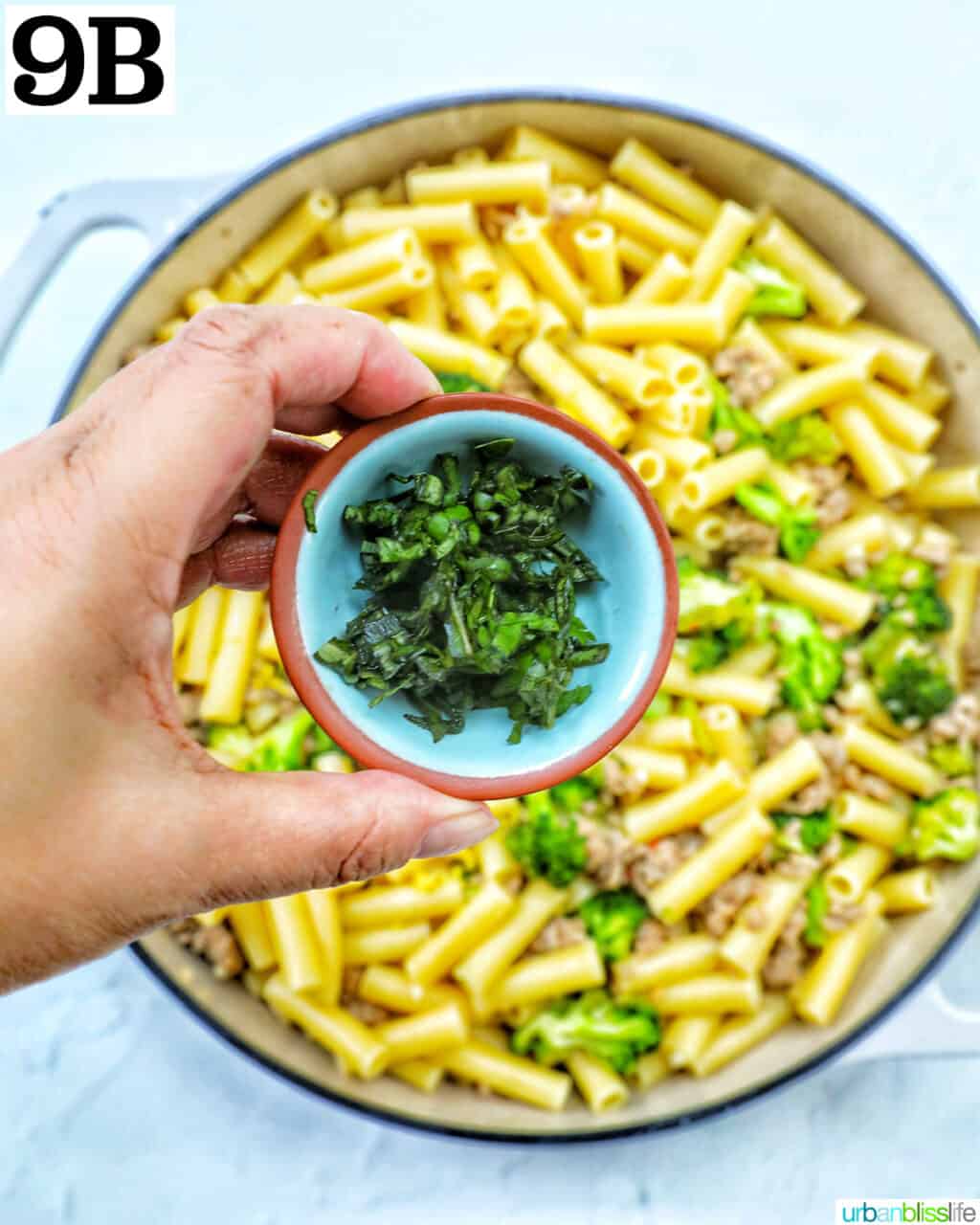 hand holding bowl of chopped herbs over a skillet of broccoli and Italian sausage.