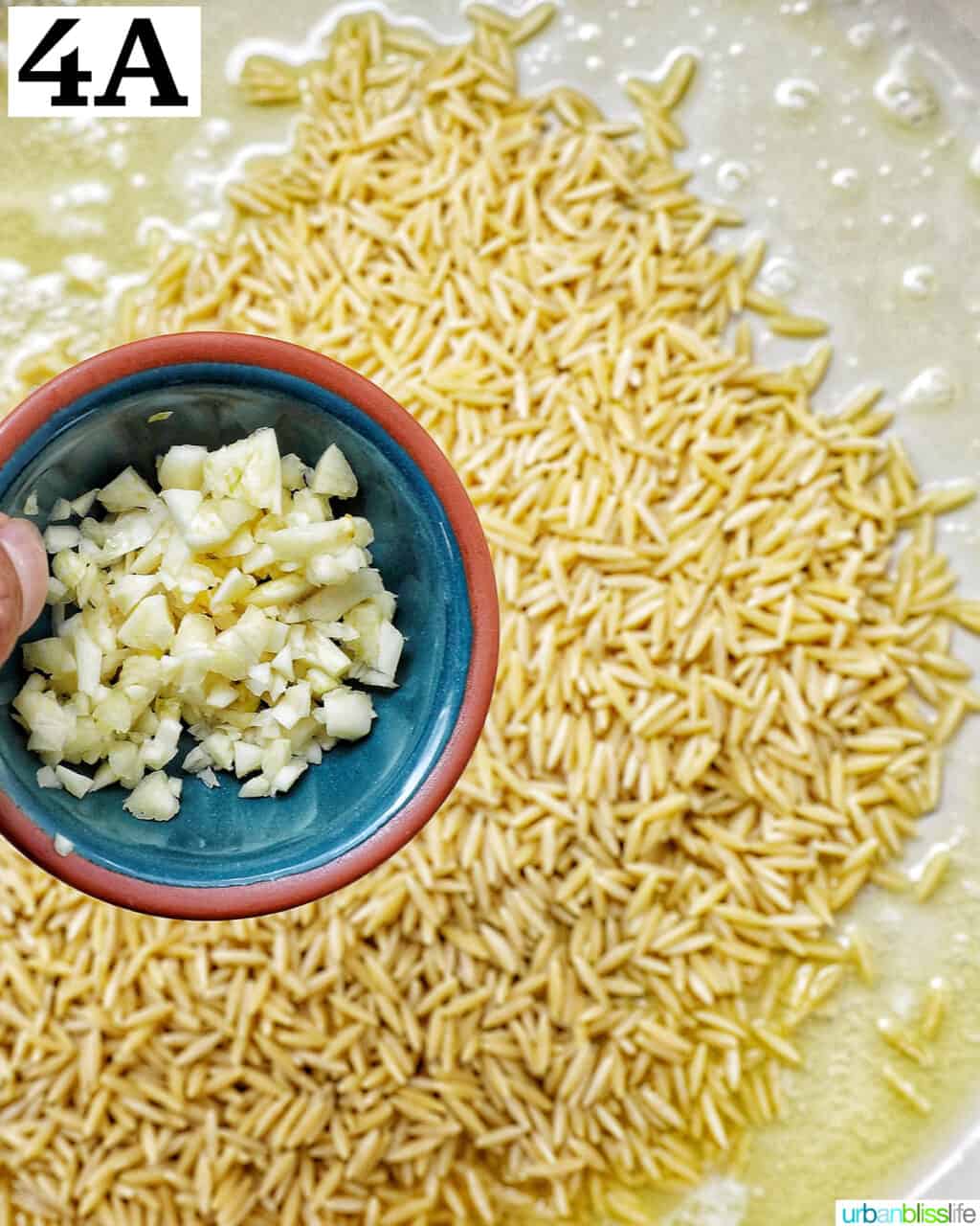 small bowl of minced garlic over a skillet with uncooked orzo.