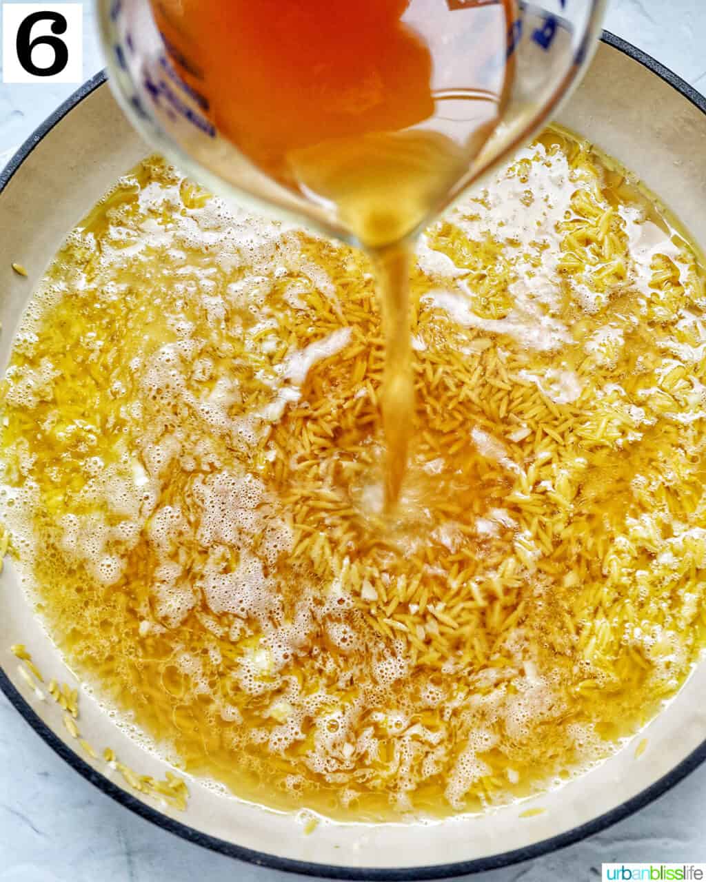 a measuring cup pouring shrimp stock into a large skillet of orzo.