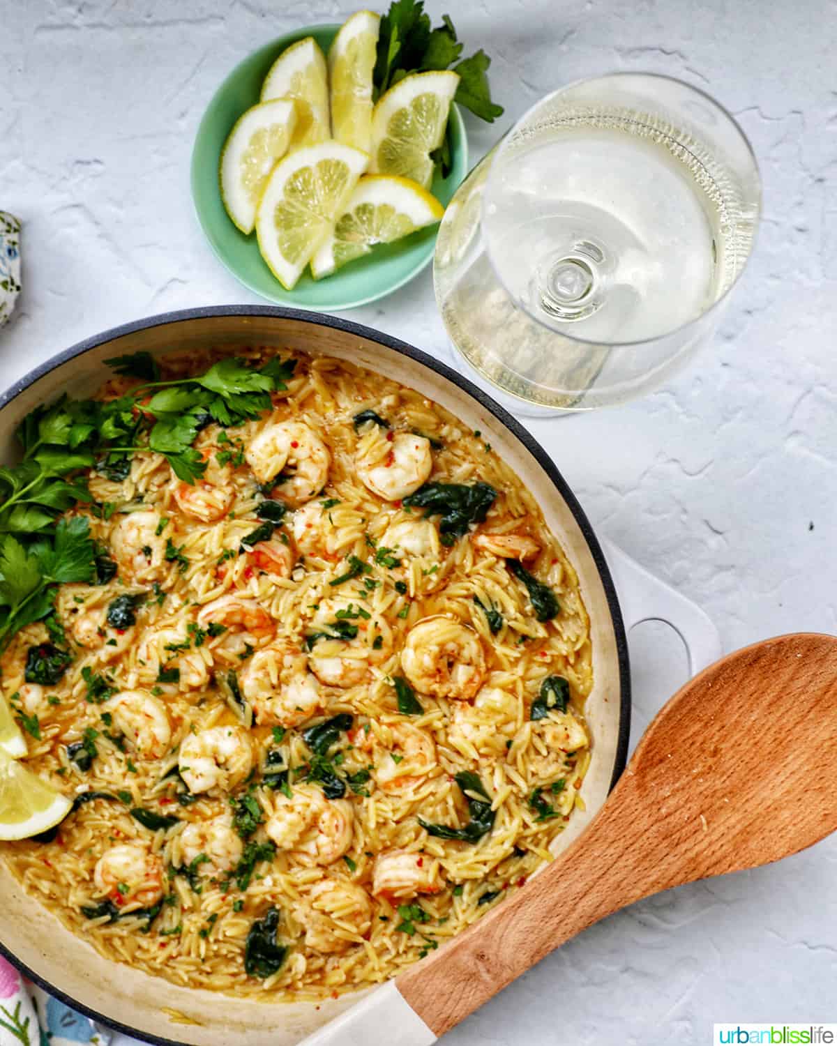 Skillet with shrimp scampi and orzo, lemons, and parsley, with a glass of white wine behind it.