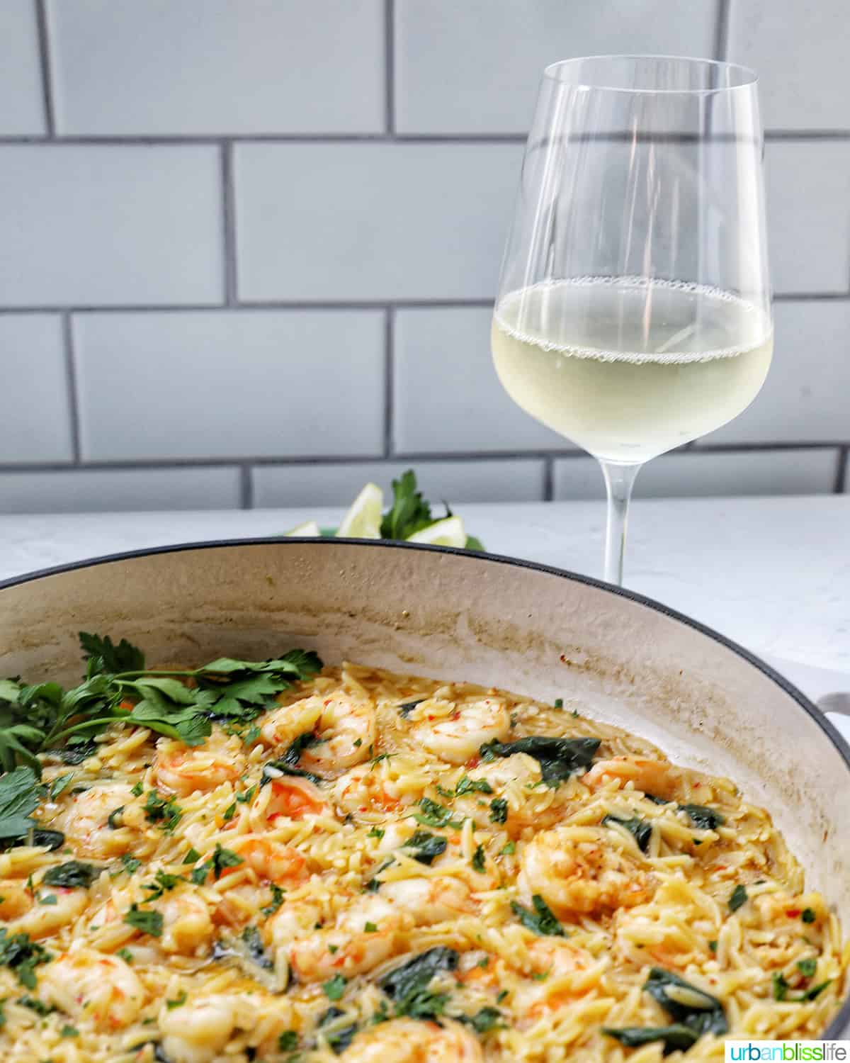 Skillet with shrimp scampi and orzo, lemons, and parsley, with a glass of white wine behind it.