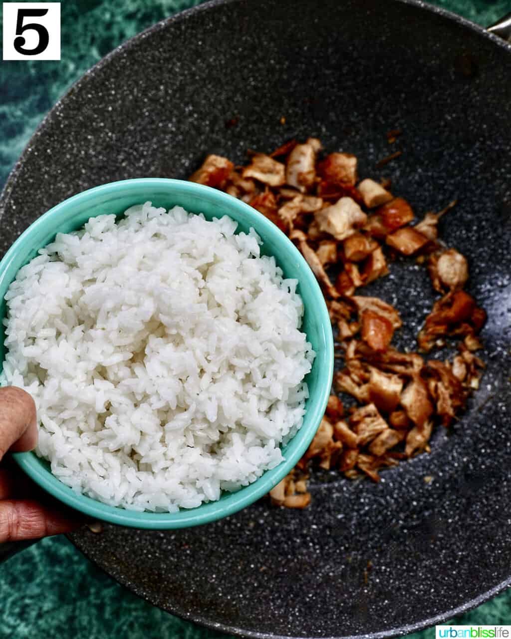 hand holding a bowl of rice over a wok of adobo chicken.