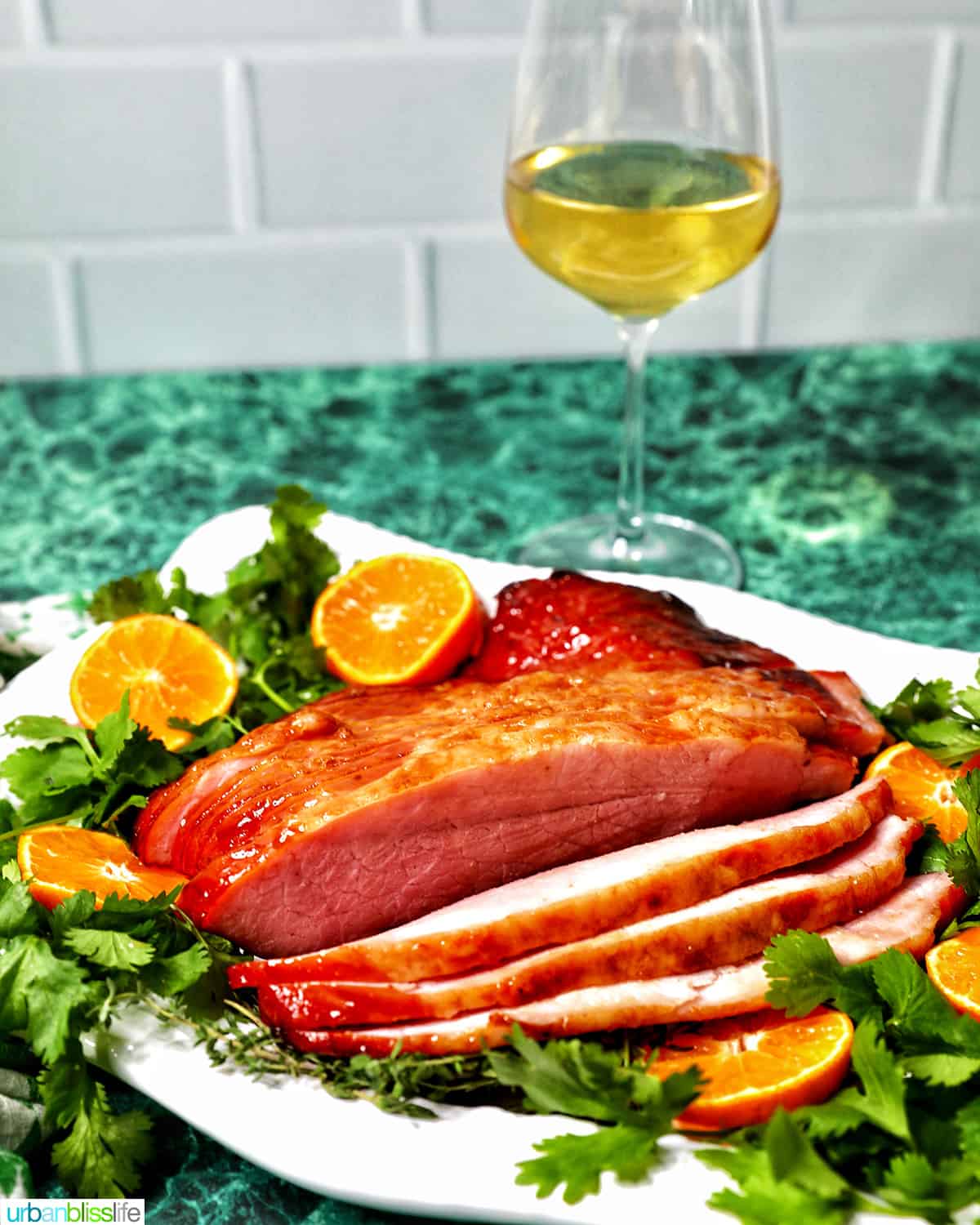 Glass of white wine behind a platter of honey glazed ham with garnishes.