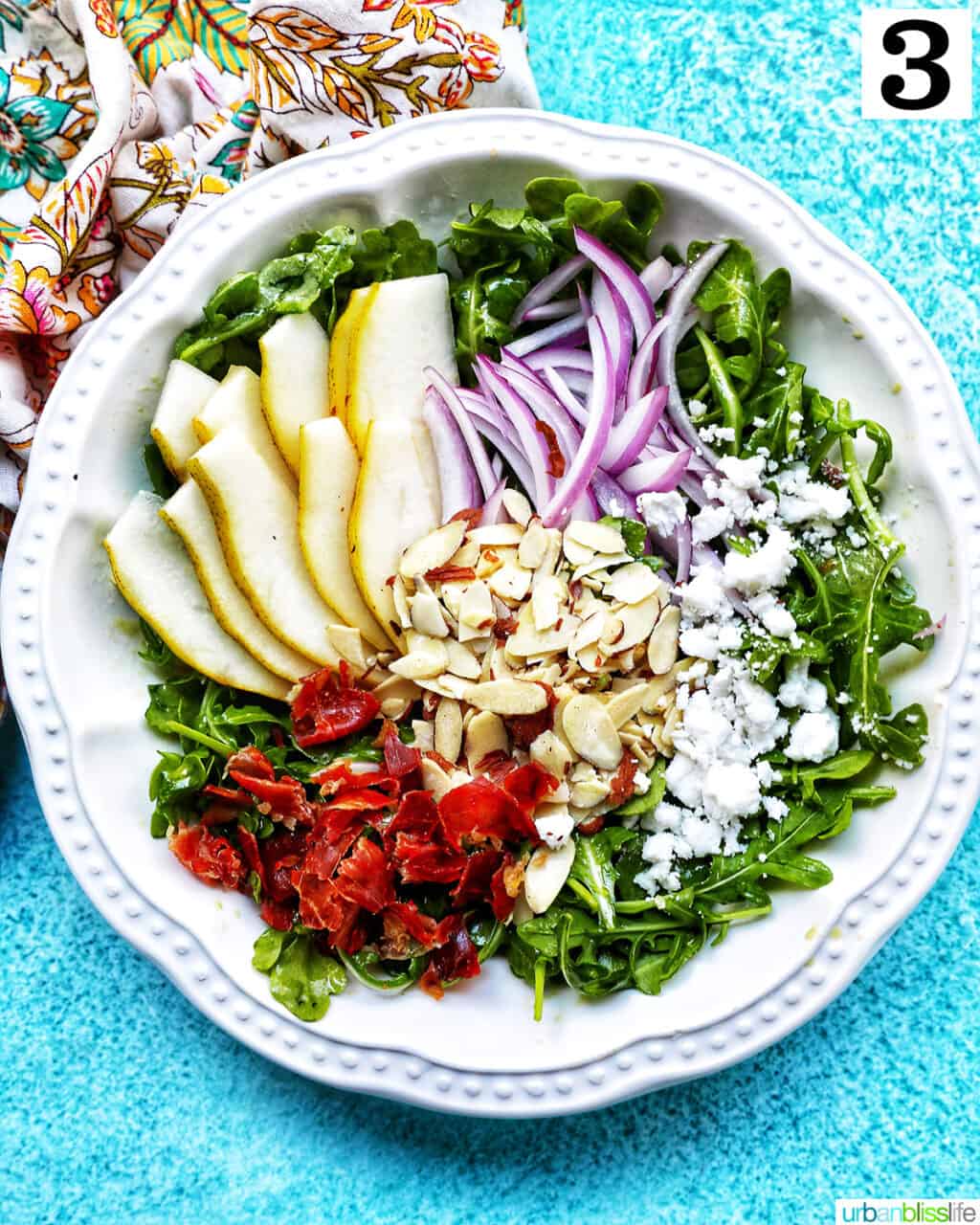 White bowl of arugula pear salad with red onions, dried cranberries, sliced almonds, and dairy-free feta cheese.