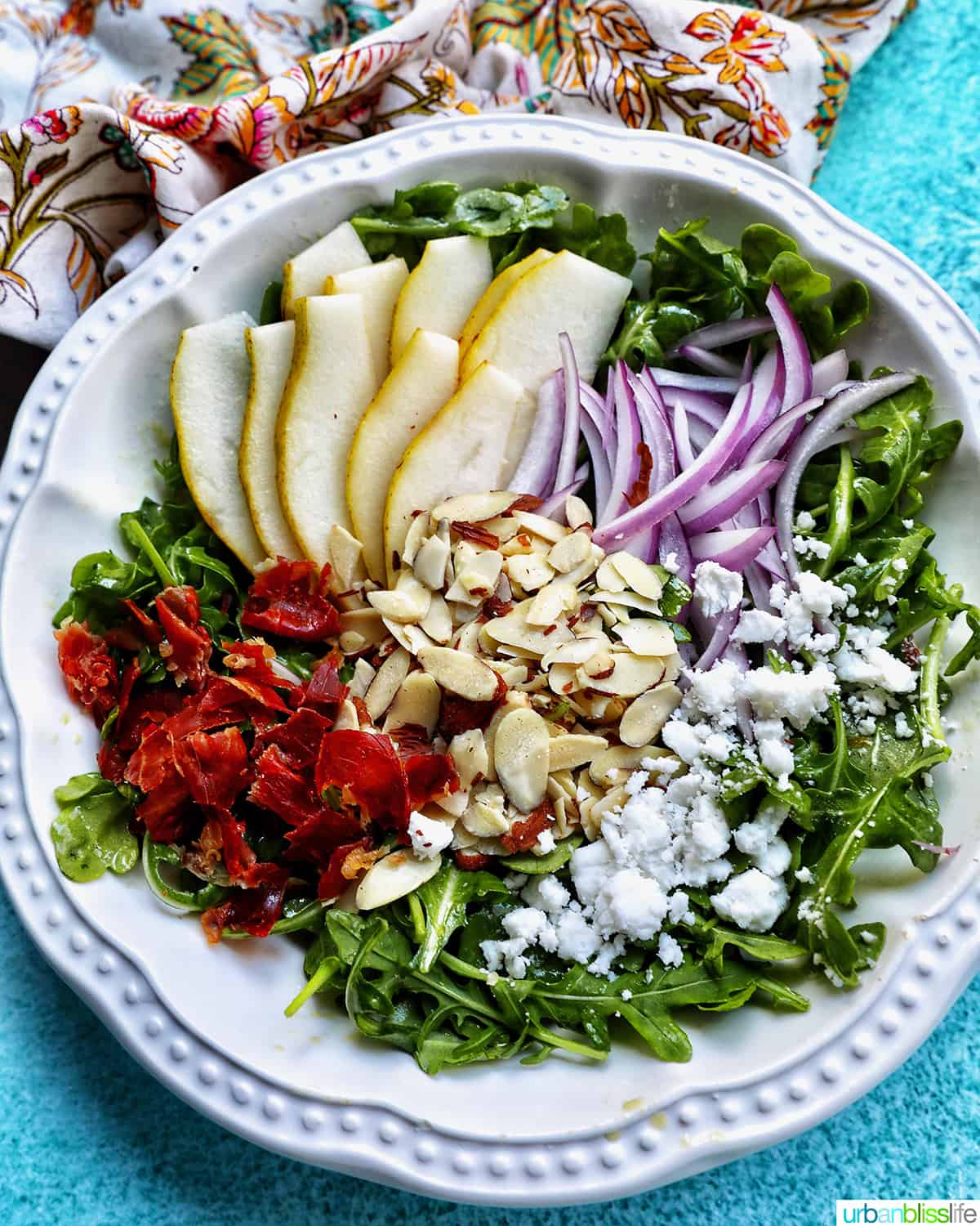 White bowl of arugula pear salad with red onions, dried cranberries, sliced almonds, and dairy-free feta cheese.