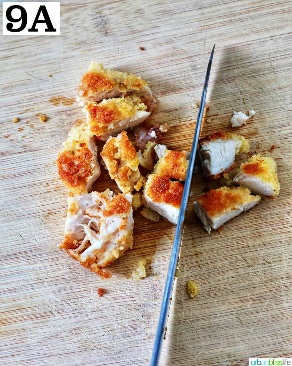 knife cutting chicken tenders into small pieces on a wooden cutting board.