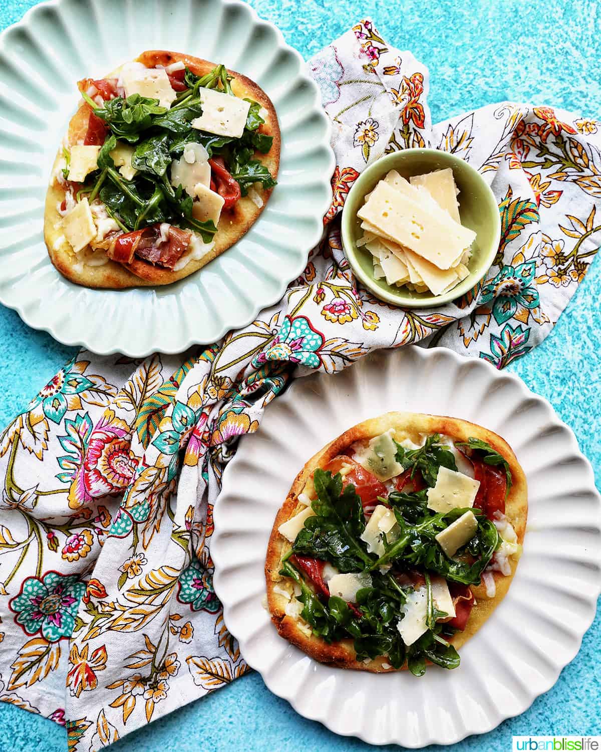 two prosciutto flatbreads with arugula and parmesan cheese on scalloped plates.