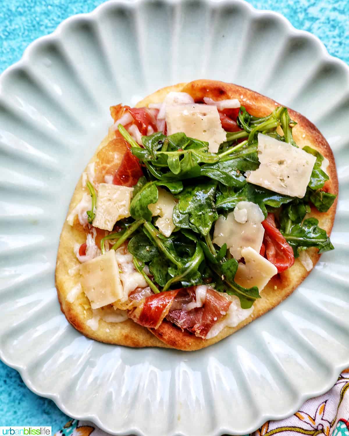 prosciutto flatbread on a scalloped plate with arugula greens and cheese.