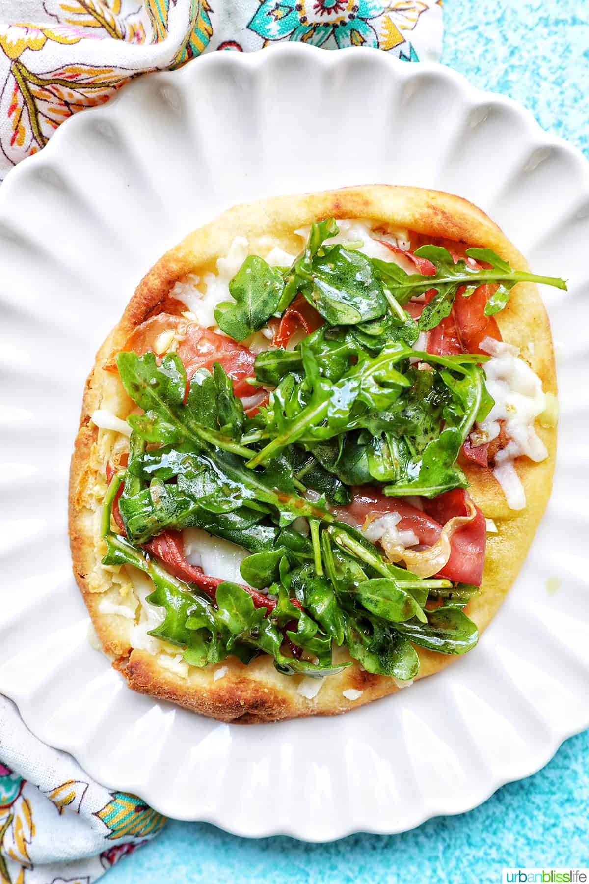 prosciutto flatbread with arugula and parmesan cheese on a scalloped plate.