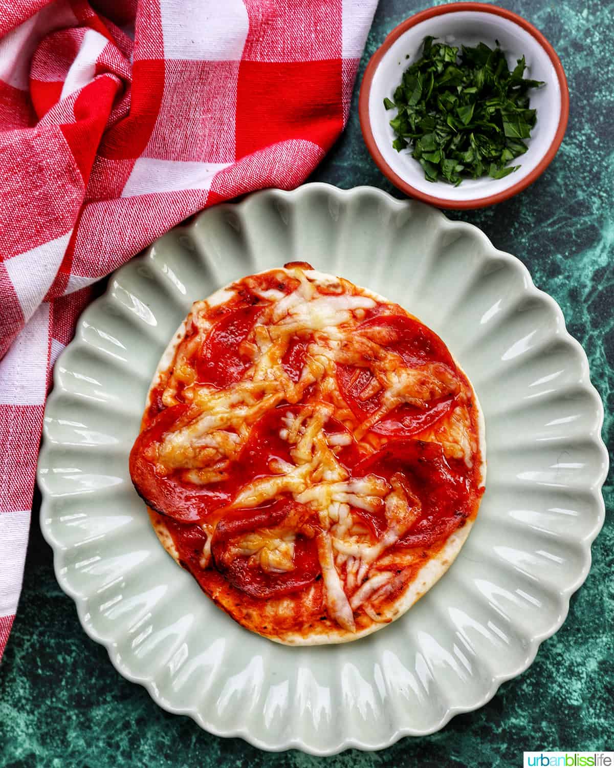 single air fryer mini pepperoni pizza on a scalloped plate next to a side of chopped fresh parsley and red and white checkered napkin.