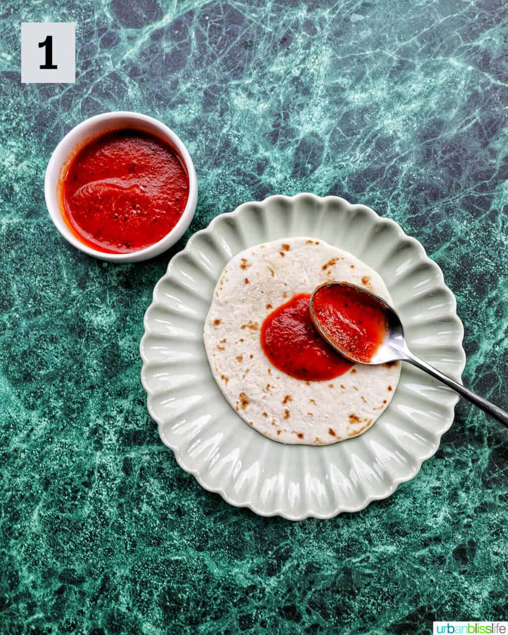 bowl of pizza sauce next to a plate with a flour tortilla with a spoon of pizza sauce on top.