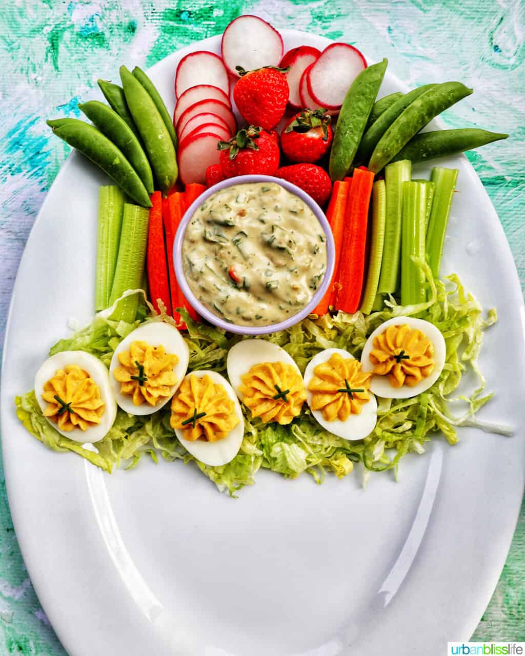 deviled eggs on shredded lettuce next to sliced vegetables and a bowl of vegetable dip on a large white platter.