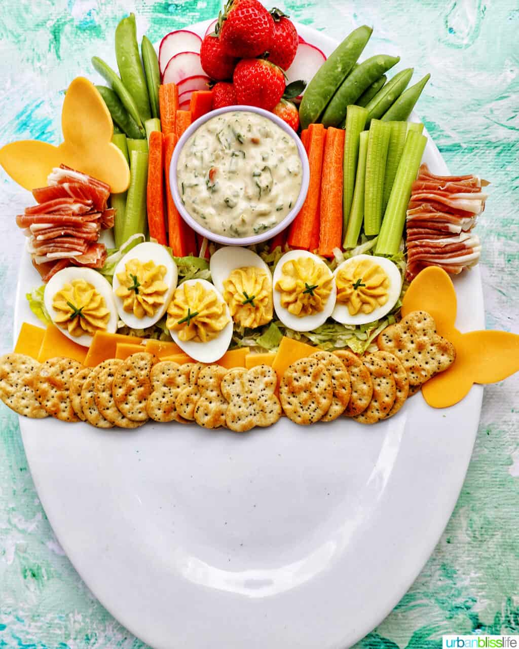 crackers, deviled eggs on shredded lettuce next to sliced vegetables and a bowl of vegetable dip on a large white platter.